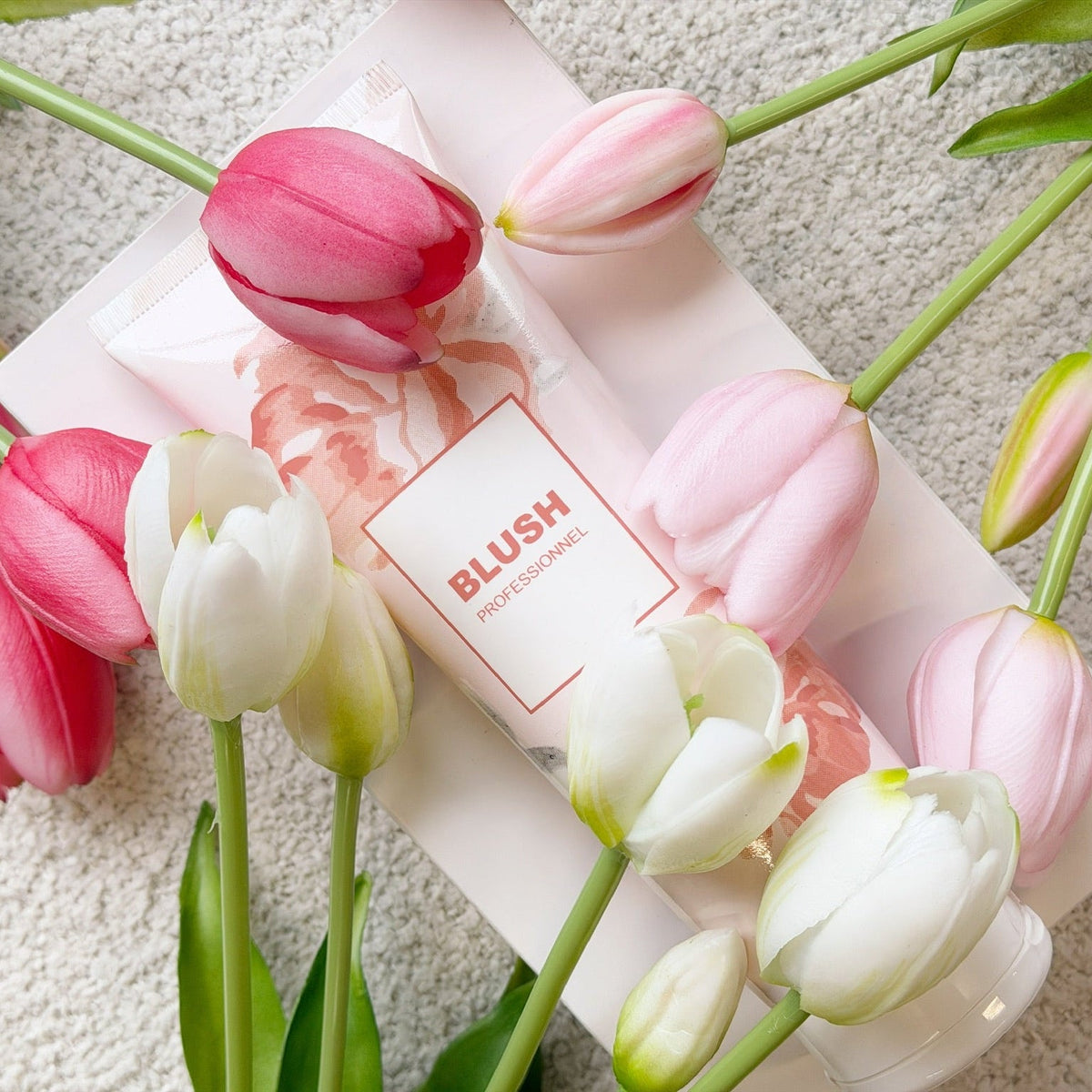 Tube de masque capillaire BLUSH posé sur une boîte rose, entouré de tulipes roses et blanches.